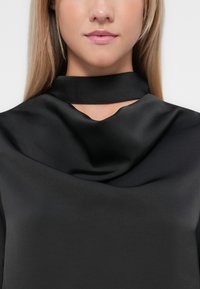 Juoda satino bluzonė su drapiruotu iškirpte ir aukštu apykakle, pasižyminti lygiu tekstūru ir švelniu blizgesiu.