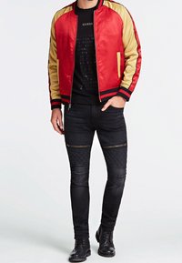 Man in een rode en gouden bomberjack, zwarte graphic t-shirt, zwarte gewatteerde ritsbroek en zwarte veterschoenen, staand met één hand in zijn zak.