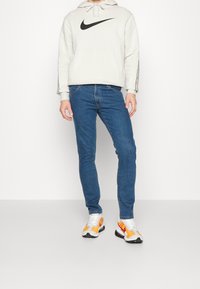 Huppari vaaleanharmaana, jossa on musta Nike-logo, siniset kapeat farkut ja moniväriset urheilukengät, joissa on valkoisia, oransseja ja mustia yksityiskohtia.