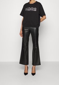 Persona vestida con una blusa de manga corta negra con "KORS" en cristales, pantalones de cuero negros acampanados y tacones altos negros puntiagudos, de pie.