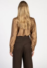 Bruine metallic blouse met lange mouwen en opstaande schouders, gecombineerd met hoge bruine geruite broek met achterzakken en taps toelopende benen.
