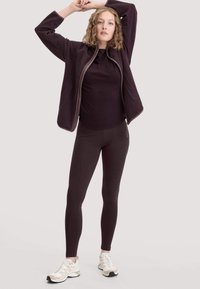 Paarse fleece jas, zwarte aansluitende top en donkere leggings. Zachte textuur met een ontspannen pasvorm. Lichtgewicht sneakers in een wit en beige kleurenschema.