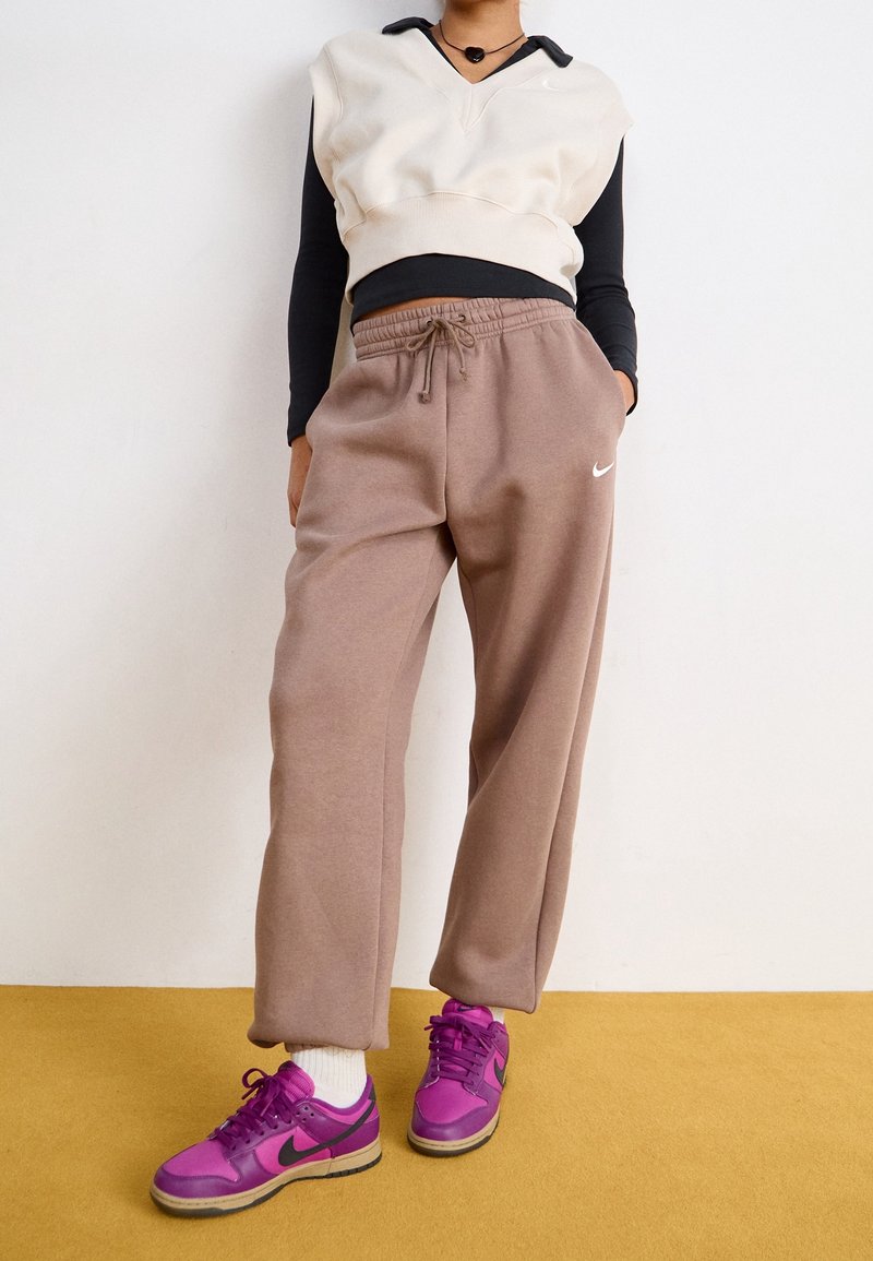 Une personne se tient contre un mur, portant un pull crème sans manches, une chemise noire, un pantalon de survêtement Nike taupe et des baskets Nike rose vif.