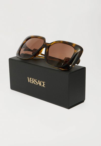 Occhiali da sole quadrati oversized in tartaruga con lenti marroni appoggiati su una scatola nera Versace con logo dorato su uno sfondo bianco.