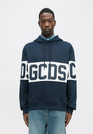 Man wearing navy blue hoodie with white band and "GCDS" text, paired with light blue jeans, standing against a plain light background.