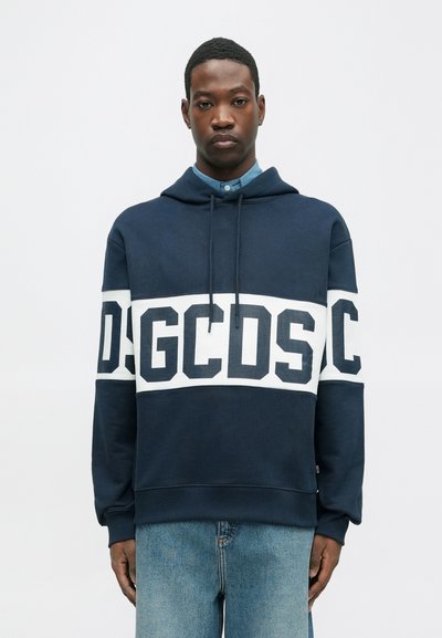 Homme portant un sweatshirt bleu marine avec une bande blanche et le texte "GCDS", associé à un jean bleu clair, se tenant devant un fond clair uniforme.