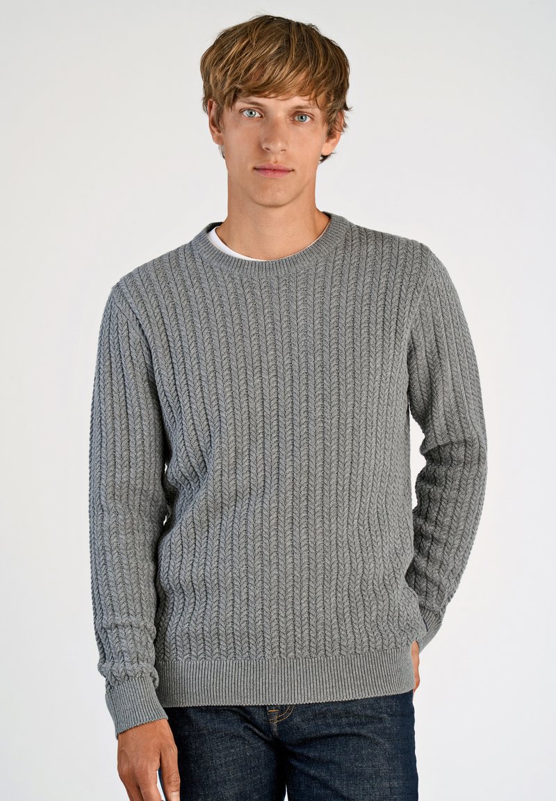 Szary sweter z dzianiny o teksturowanym, kablowym wzorze. Okrągły dekolt oraz ściągacze przy mankietach i u dołu. Noszony na białej koszuli, w zestawieniu z ciemnymi dżinsami.