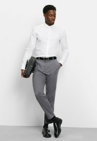 Weißes langärmeliges Hemd, graue maßgeschneiderte Hose mit Gürtel, schwarze Dressschuhe und schwarze Umhängetasche. Gepflegtes, professionelles Erscheinungsbild.