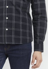 Camisa de cuadros azul marino en tejido de algodón con líneas a cuadros blancas, con botones en la parte delantera y mangas largas, con puños en las muñecas.
