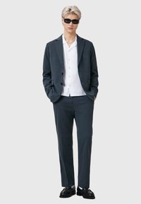 Mörkgrå kostym med slim fit, i kombination med en vit skjorta och svarta loafers. Modellen bär mörka solglasögon och har ljust hår.