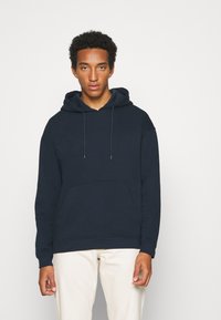 Marineblaue Baumwollkapuzenjacke mit Kordelzügen, einer vorderen Kängurutasche und gerippten Bündchen. Getragen mit hellen Hosen.