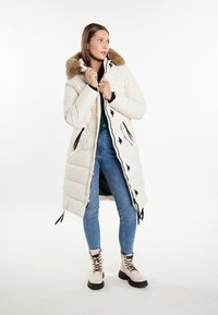 Lyscremet polstring parka med pelsforet hætte, sorte detaljer og sidelommer; parret med blå denim og beige snørestøvler.