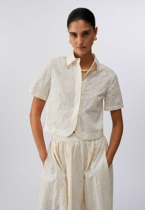 Donna con capelli scuri pettinati all'indietro indossa una camicetta crema ricamata a maniche corte e una gonna a vita alta abbinata, con le mani nelle tasche.