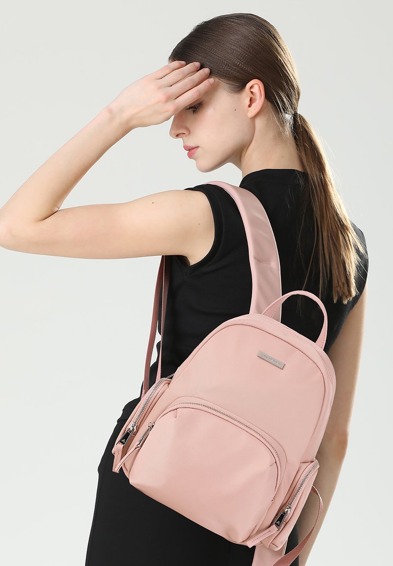 Zaino in nylon rosa chiaro con più tasche con zip, forma curva e spallacci regolabili, indossato su una spalla.