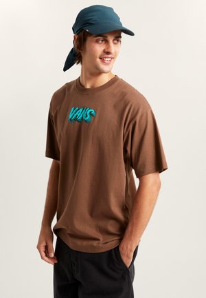 T-shirt en coton marron avec un logo "VANS" turquoise, manches courtes, col rond, coupe décontractée. Porté avec une casquette bleue dotée d'un rabat attaché.