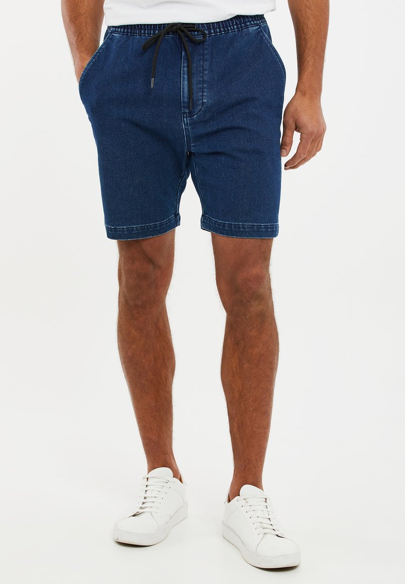 Pantalones cortos de mezclilla en azul oscuro, con una cintura elástica y cordón, bolsillos laterales y un diseño casual. Combinados con zapatillas blancas.