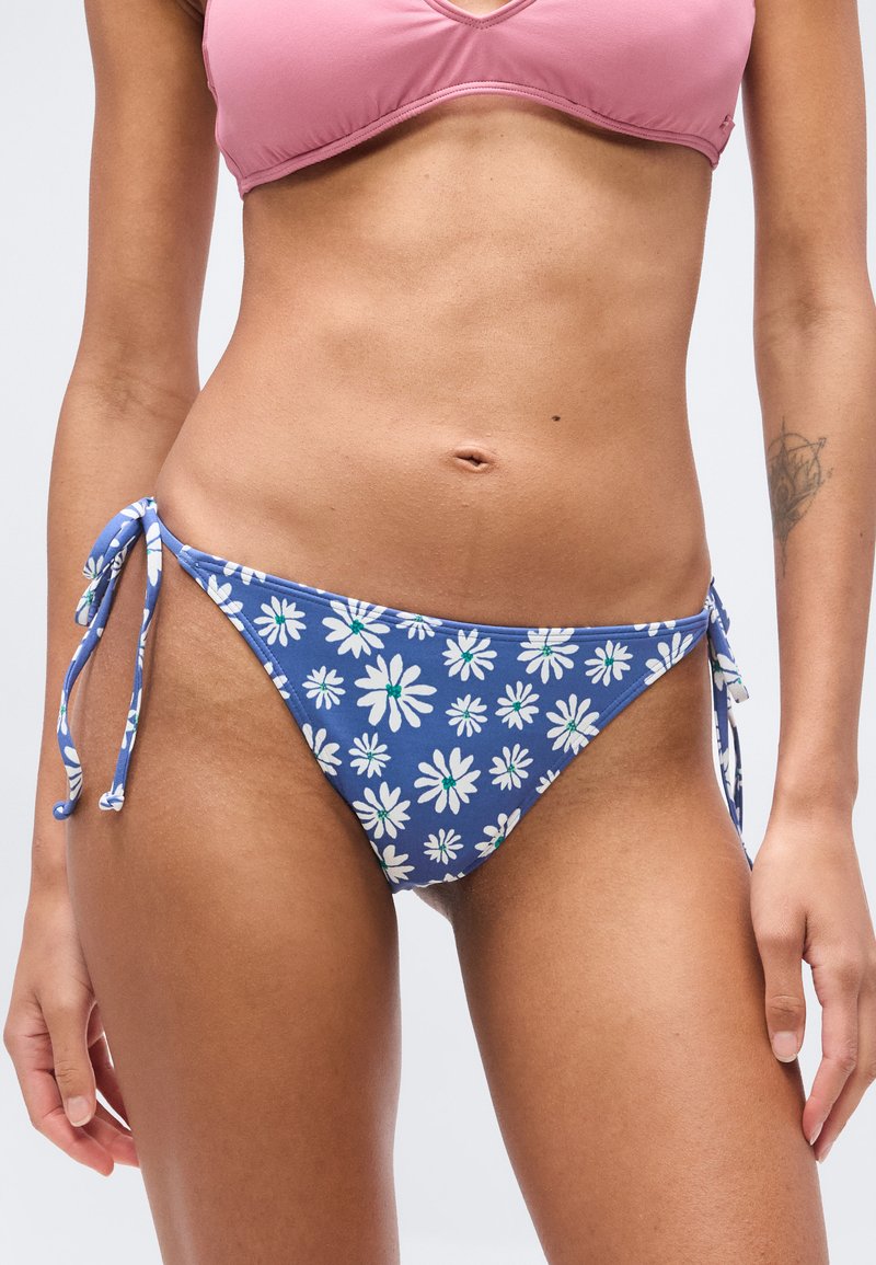 Femme portant un haut de bikini rose et un bas de bikini bleu à motifs floraux avec des liens sur les côtés, torse et jambes supérieures visibles, tatouage sur l'avant-bras gauche.