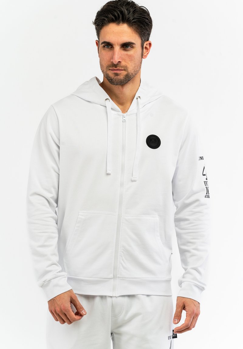 Sudadera blanca con cremallera y capucha ajustable, bolsillos frontales y un parche negro con logo, con un sutil detalle de texto en las mangas.