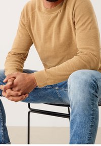 Beżowy sweter dzianinowy z prążkowanymi mankietami i okrągłym dekoltem, zestawiony z jasnoniebieskimi dżinsami. Siedzi na czarnej metalowej krześle.