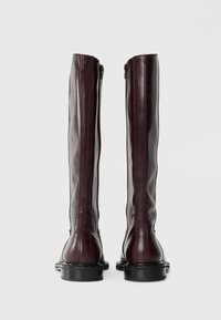Bottes en cuir bourgogne jusqu'aux genoux avec une finition lisse, dotées d'une fermeture éclair à l'arrière et d'une semelle en caoutchouc noire contrastante. Coutures visibles.