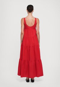 Abito maxi rosso senza maniche con schiena bassa, design a strati e tessuto fluido. Stilizzato con orlo semplice e indossato con scarpe nere.