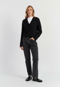 Cardigan nero a maglia con scollo a V, chiusura con bottoni e orlo a costine, abbinato a jeans grigi scuri a gamba dritta e scarpe slip-on nere.