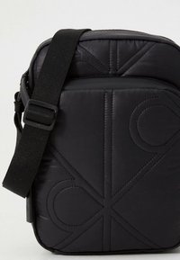 Black padded shoulder bag with a textured surface, featuring geometric stitching and an adjustable strap. Compact rectangular shape.