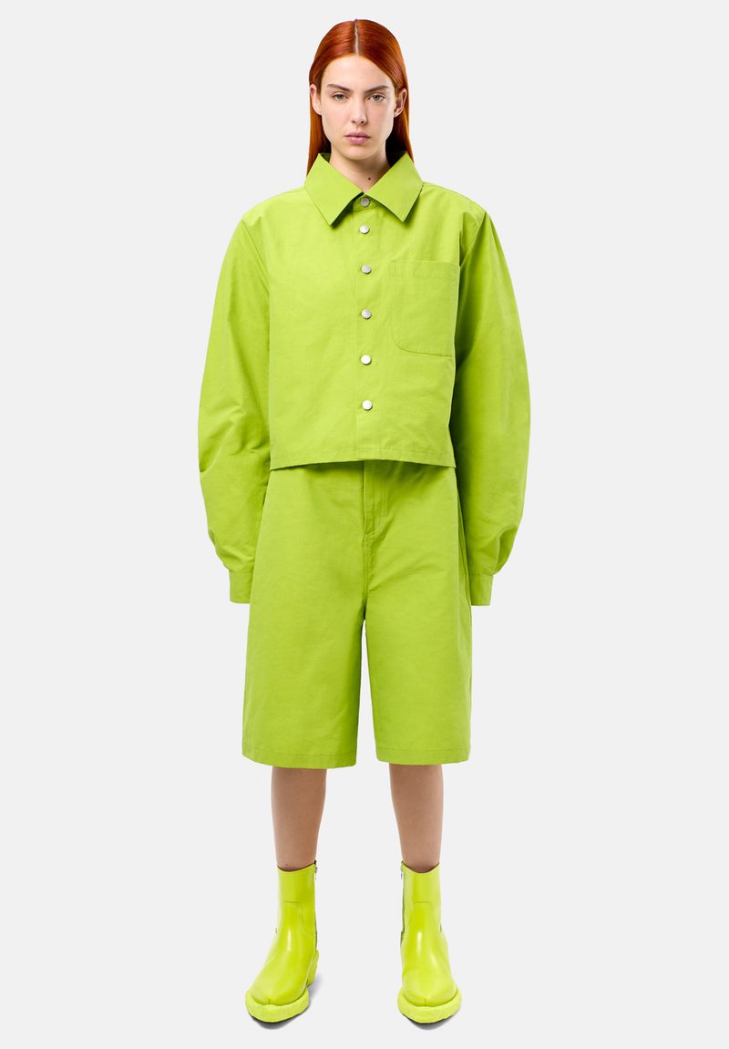 Zkrácená lime-green bunda s velkým límcem a stříbrnými knoflíky, v kombinaci s odpovídajícími šortkami a lesklými lime-green kotníkovými botami.
