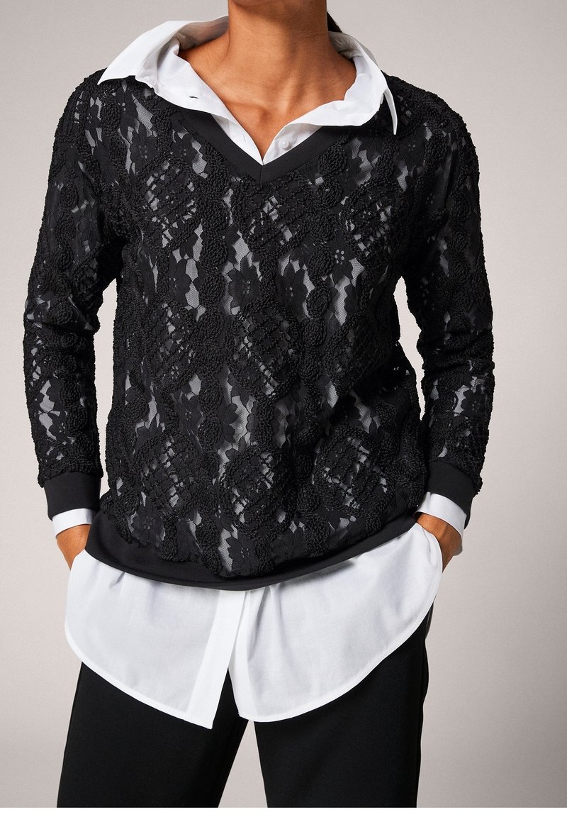 Personne portant un haut à manches longues noir à motif en dentelle par-dessus une chemise blanche à col, les mains dans les poches, habillée d'un pantalon noir.