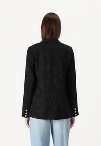 Blazer noir texturé avec un col, des manches longues et trois accents de boutons en argent sur chaque poignet. Porté sur un pantalon bleu clair.
