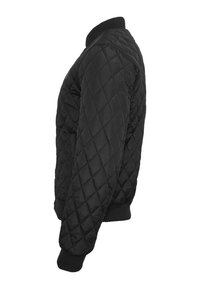 Veste matelassée noire avec couture en forme de losange, poignets, ourlet et col côtelés, vue de profil sur fond blanc.