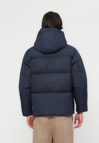 Donkerblauwe gewatteerde jas met capuchon, met een doorgestikt ontwerp, elastische manchetten en een rechte snit, weergegeven vanaf de achterkant.