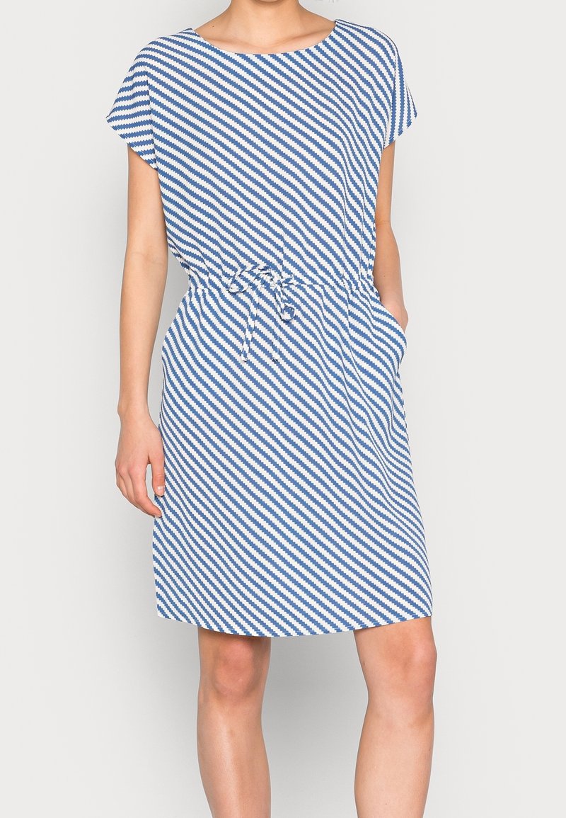 Femme portant une robe bleu et blanc rayée en diagonale au genou, manches courtes et ceinture à la taille, une main dans la poche.