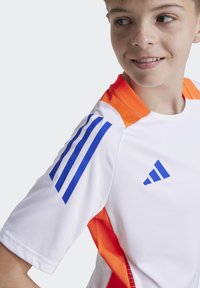 Fehér sportpóló narancssárga díszítéssel, kék csíkozás van a vállakon és egy kék logó. Simább anyagú, rövid ujjú.
