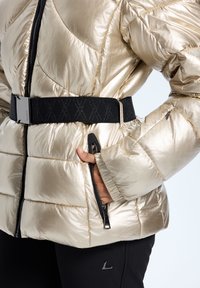 Metallisk guld pufferjakke med diagonal quiltet tekstur, sort bælte med logo og sidelommer med lynlås. Blødt finish, struktureret silhuet.