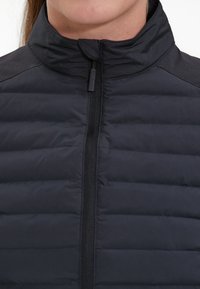 Gilet imbottito nero con colletto alto, design a righe orizzontali e una zip frontale con un piccolo tirante nero.