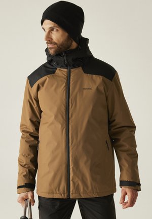 Hombre con chaqueta con capucha marrón y negra y gorro negro mirando hacia la izquierda, sosteniendo bastones de trekking en una mano contra un fondo liso.