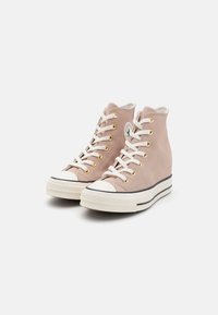 Baskets montantes en suède rose pâle avec des capuchons en caoutchouc blanc, des œillets dorés, des lacets crème et une semelle à plateforme avec des rayures noires.