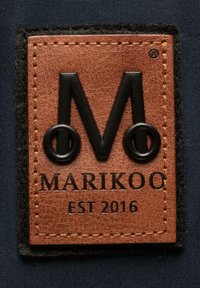 Patch en cuir brun avec un logo "M" noir et le texte "MARIKOO EST 2016" cousu sur un tissu foncé.
