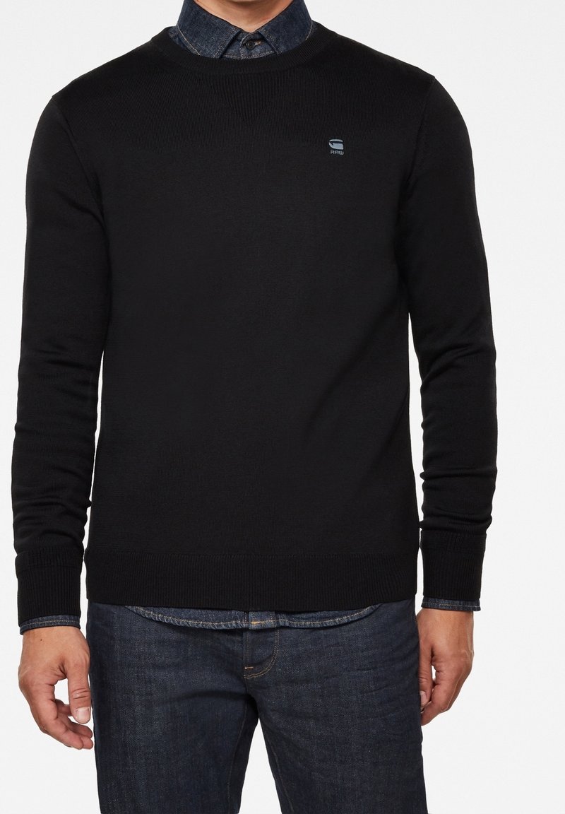 Schwarzer Strickpullover mit langen Ärmeln und einem gerippten Saum. Verfügt über ein Logo auf der Brust und wird mit einem Jeanshemd darunter kombiniert.