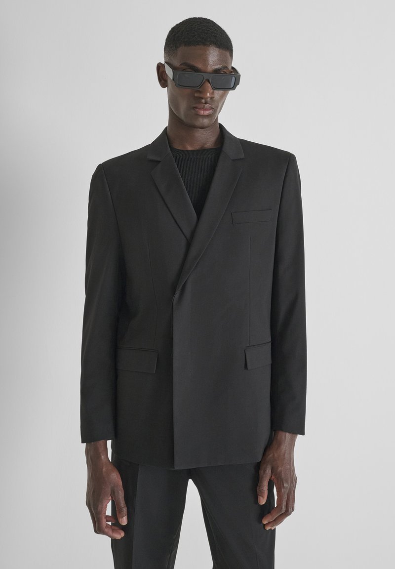 Crni sako krojenog izgleda s reverima, jednim džepom na prsima i dva prednja džepa. Glatka tkanina s strukturiranim krojem.