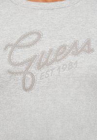 Gray crewneck sweatshirt featuring "Guess" in a cursive design with silver studs and "EST. 1981" embossed below. Smooth fabric texture.