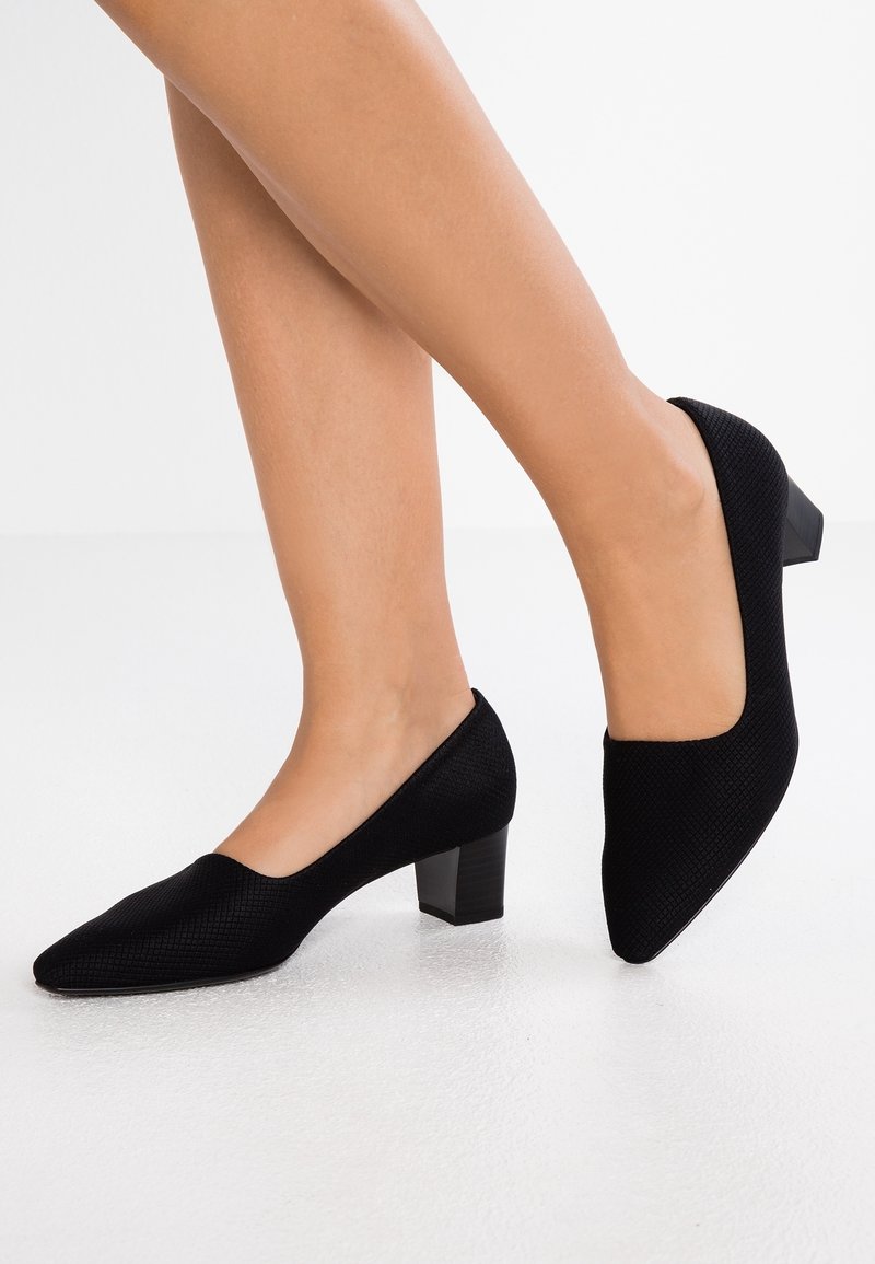 Jambes de femme portant des chaussures noires texturées à talons bas et bouts pointus sur un sol blanc.