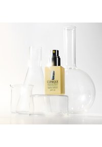 Clinique fuktighetskremflaske i gult glass med svart pumpe, merket "SPF 50", plassert blant klare glasslaboratoriekolber på en hvit bakgrunn.