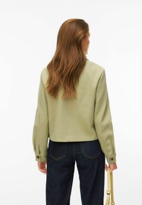 Femme aux longs cheveux bruns vue de dos, portant une veste vert clair, un jean foncé, et tenant un sac à main jaune à ses côtés.