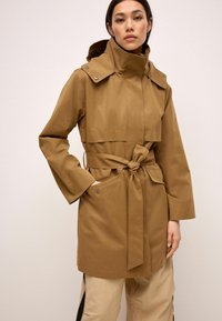 Femme portant un trench-coat ceinturé de couleur marron clair avec un col montant, se tenant avec une main dans la poche du manteau devant un fond uni.