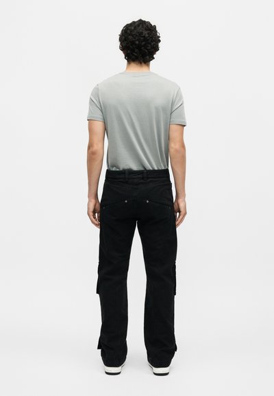 Άντρας με σγουρά μαλλιά που φοράει ανοιχτογκρίζο κοντομάνικο πουκάμισο, μαύρο παντελόνι cargo και λευκά αθλητικά, στέκεται γυρισμένος προς τα πίσω.