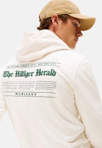 Joven con gorra beige y sudadera blanca con texto verde "The Hilfiger Herald" y un párrafo en la parte de atrás, mirando hacia la derecha.