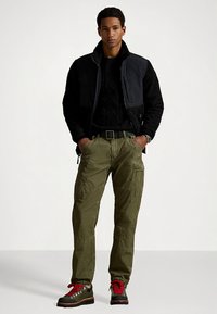 Μαύρο, υφασμάτινο σακάκι πάνω από μαύρο πλεκτό πουλόβερ, ελιάς παντελόνι cargo και μποτάκια πεζοπορίας με κόκκινες λεπτομέρειες. Το μοντέλο στέκεται με τα χέρια στις τσέπες.