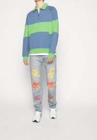 Pull-over bicolore bleu et vert, col zippé, associé à un jean en denim clair orné de motifs colorés de fleurs et d'yeux. Baskets blanches.